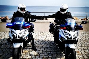 Na zdjęciu dwaj policjanci na motocyklach, którzy znajdują się na pasie nadmorskim. Za nimi w tle woda