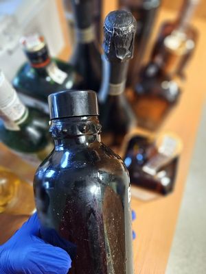 Na zdjęciu butelka alkoholu, która trzymana jest w dłoni z założoną niebieską rękawiczką. W tle inne butelki alkoholu.