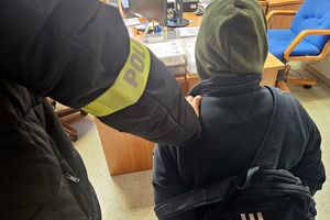 Na zdjęciu zatrzymany mężczyzna, który siedzi na krześle przed biurkiem. Obok niego fragment sylwetki policjantka w stroju cywilnym i z założoną na ramieniu opaską z napisem POLICJA.