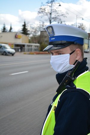 na zbliżeniu od pasa w górę umundurowany policjant, który patrzy na ulicę. Za nim widoczna ulica i sklep