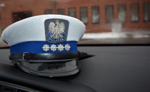 Wnętrze radiowozu, gdzie na podszybiu auta leży czapka policjanta ruchu drogowego w stopniu aspiranta sztabowego. Przez szybę przednią auta widać ścianę budynku o kratowanymi oknami