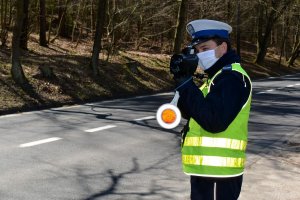 Umundurowany policjant trzymając urządzenie do pomiaru prędkości mierzy prędkość pojazdów stojąc przy ulicy. Za policjantem widać las.