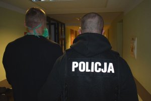 Korytarz. Policjant w stroju cywilnym z kamizelką taktyczną z napisem POLICJA prowadzi zatrzymanego mężczyznę. Obu mężczyzn widać od pasa w górę