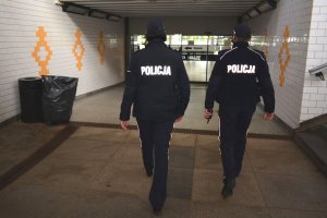 Tunel będący ciągiem pieszych. Umundurowani policjanci idą tunelem w stronę pasażu. W oddali za szklanymi drzwiami widać niewyraźnie sylwetki osób