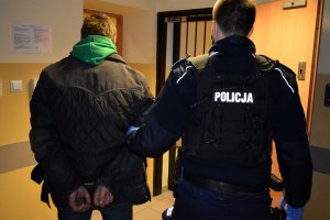 Korytarz w Komendzie Miejskiej Policji w Sopocie. Umundurowany policjant prowadzi zatrzymanego mężczyznę, który ma ręce z tyłu skute kajdankami