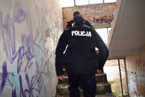 Wnętrze pustostanu. Umundurowani policjanci idą po schodach do góry. Na ścianach widoczne graffiti i tagi.