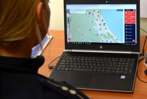 Pomieszczenie biurowe. Widać fragment sylwetki umundurowanego policjanta, który siedzi przy biurku. Na biurku leży laptop, na którym wyświetlona jest strona Krajowej Mapy Zagrożeń Bezpieczeństwa. Policjant patrzy się w laptopa.