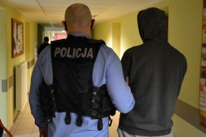 Korytarz. Policjant w stroju cywilnym z nałożoną kamizelką z napisem POLICJA prowadzi zatrzymanego mężczyznę. Za nimi w tle widać fragment sylwetki innej  osoby.