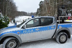 Widok na ośnieżony stok narciarski, przed którym stoi oznakowany policyjny radiowóz. Na stoku widoczne sylwetki osób.