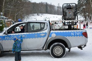 Widok na ośnieżony stok narciarski, przed którym stoi oznakowany policyjny radiowóz. Przed stokiem stoi małe dziecko i rozmawia z policjantem. Na stoku widoczne sylwetki osób.