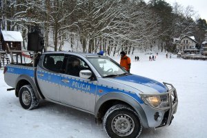 Widok na ośnieżony teren. Stoi oznakowany policyjny radiowóz, w którym siedzą policjanci. Jeden z nich rozmawia z mężczyzną stojącym za radiowozem. Za radiowozem widoczne drzewa.