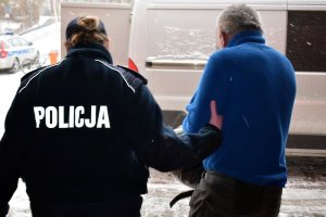 Umundurowana policjantka prowadzi zatrzymanego mężczyznę. W tle widoczny samochód typu bus i oznakowany radiowóz. Na ziemi leży śnieg.