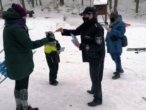 Przestrzeń otwarta, przy lesie. Umundurowana policjantka wręcza ulotkę kobiecie. Na drugim planie dzieci i osoby dorosłe.