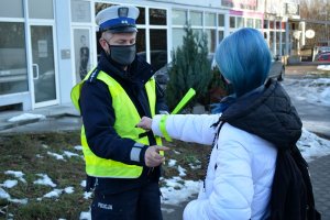 Umundurowany policjant wręcza na chodniku odblask kobiecie ubranej w białą kurtkę. Za nimi widać fragment ulicy i budynku.