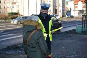 Umundurowany policjant wręcza odblask kobiecie. Osoby stoją na chodniku. Za nimi widać ulicę i budynki.