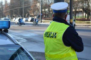 Umundurowany policjant stoi w okolicy skrzyżowania ulic i przejść dla pieszych przez ulicę. Policjant obserwuje osoby widoczne w oddali, któe przechodzą przez przejście dla pieszych. Za nimi na ulicy stoją samochody.