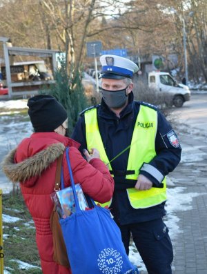 Umundurowany policjant wręcza odblask kobiecie. Osoby stoją na chodniku. Za nimi widać ulicę.