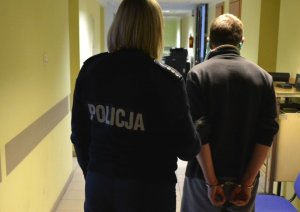 Korytarz.  Umundurowana policjantka prowadzi zatrzymanego mężczyznę, który  ma założone kajdanki na ręce trzymane z tyłu.