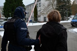 Umundurowany policjant prowadzi zatrzymanego mężczyznę. W tle widoczne oznakowane radiowozy.