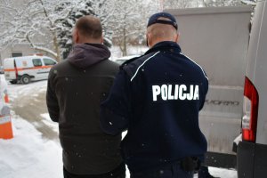 Umundurowany policjant prowadzi zatrzymanego mężczyznę do nieoznakowanego radiowozu