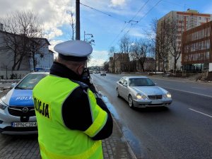 Umundurowany policjant trzymając w ręku urządzenie mierzy prędkość jadących pojazdów. Policjant stoi przy jezdni, po której jadą samochody. Za policjantem widoczny fragment radiowozu i budynki znajdujące się przy ulicy