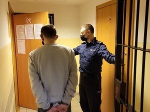 Korytarz w budynku. Umundurowany policjant wprowadza do pomieszczenia zatrzymanego mężczyznę, który ma założone kajdanki na ręce trzymane z tyłu