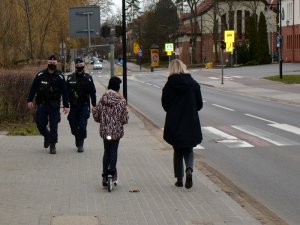 Umundurowani policjanci idą chodnikiem wzdłuż ulicy. Przed nimi dwie inne osoby idą chodnikiem w przeciwnym kierunku. W tle widać jadące ulicą samochody