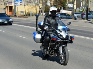 Fragment ulicy. Policjant siedząc na oznakowanym motocyklu jedzie ulicą.