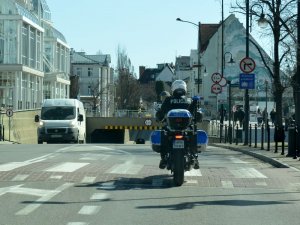 Fragment ulicy. Policjant siedząc na oznakowanym motocyklu jedzie ulicą. Po bokach widać budynki, chodniki i przechodniów.