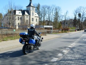 Fragment ulicy. Policjant siedząc na oznakowanym motocyklu jedzie ulicą.