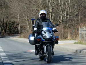 Fragment ulicy. Policjant siedząc na oznakowanym motocyklu jedzie ulicą.