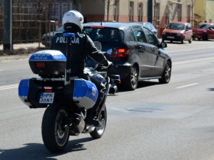 Fragment ulicy. Policjant siedząc na oznakowanym motocyklu jedzie ulicą. Za nim widoczne pojazdy.