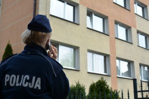 Umundurowana policjantka trzyma w ręku telefon i patrzy się w stronę okien budynku