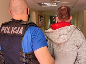 Policjant w stroju cywilnym i z założoną kamizelką z napisem POLICJA prowadzi korytarzem zatrzymanego mężczyznę. Sylwetki osób są widoczne od pasa do głowy.