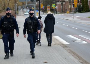 Umundurowani policjanci idą chodnikiem wzdłuż ulicy. Za nimi widoczni piesi.