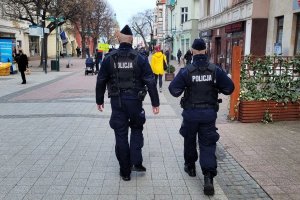 Umundurowani policjanci idą deptakiem dla pieszych. Obok idą inne osoby.