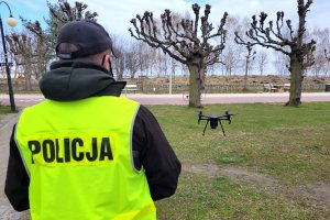 Na zdjęciu policjant w stroju cywilnym i z założoną kamizelką z napisem POLICJA, który stoi w parku i obsługuje drona widocznego za nim