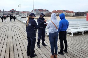 Umundurowane policjantki przeprowadzają interwencję wobec dwóch osób. Osoby stoją na molo. Za nimi widoczne budynki, plaża i inne osoby.