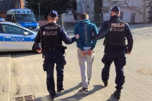 Umundurowani policjanci prowadzą do radiowozu zatrzymanego mężczyznę, który ma założone na ręce kajdanki.