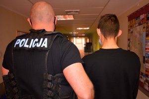 Policjant w stroju cywilnym i z założoną kamizelka z napisem POLICJA prowadzi korytarzem zatrzymanego mężczyznę