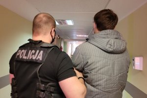 Policjant w stroju cywilnym i z założoną kamizelką z napisem POLICJA prowadzi korytarzem zatrzymanego mężczyznę