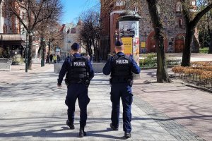 Umundurowani policjanci idą deptakiem. Za nimi w tle widoczne sylwetki innych osób.