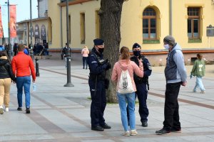 Miejsce publiczne, deptak. Umundurowani policjanci przeprowadzają interwencję z dwiema osobami. W tle inne osoby, które idą deptakiem