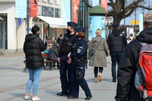 Miejsce publiczne, deptak. Umundurowani policjanci rozmawiają z kobietą. Obok nich deptakiem spacerują inne osoby.