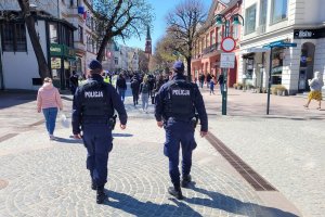 Umundurowani policjanci idą ulicą. Za nimi widoczne budynki i spacerujące osoby.