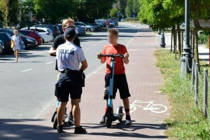 Droga dla rowerów przy pasie jezdni. Umundurowana policjantka rozmawia z mężczyzną i chłopcem, którzy trzymają elektryczne hulajnogi. W tle widoczne samochody i kobieta idąca wzdłuż zaparkowanych samochodów.