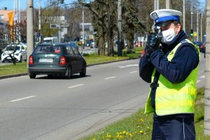 Umundurowany policjant stoi przy jezdni i za pomocą urządzenia trzymanego w rękach mierzy prędkość pojazdów. Za policjantem w tle widoczna ulica i samochody.