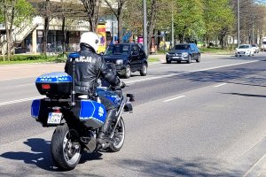 Policjant na oznakowanym motocyklu jedzie ulicą. Na przeciwległych pasach ruchu widać inne pojazdy.