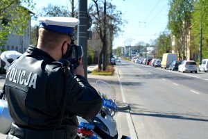 Policjant ruchu drogowego stojąc przy jezdni, po której jadą samochody, za pomocą urządzenia mierzy prędkość pojazdów. Za policjantem widać fragment oznakowanego motocykla.