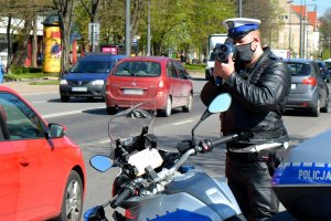 Policjant ruchu drogowego stoi przy jezdni, po której jadą samochody i za pomocą urządzenia mierzy prędkość pojazdów. Przed policjantem widać fragment oznakowanego motocykla.